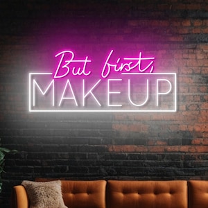 Puede incluir: Un letrero de neón rosa con borde blanco en una pared de ladrillo. El letrero dice "But first, MAKEUP".