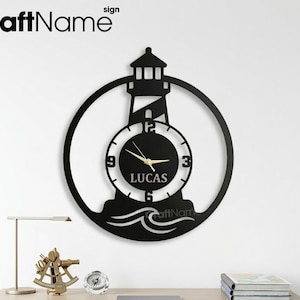 May include: Black metal lighthouse wall clock with a circular design. The clock face displays the name "LUCAS" and gold-colored hands. The clock is mounted on a white wall above a desk with decorative items.