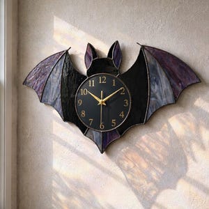 Puede incluir: Un reloj de pared con forma de murciélago con esfera negra y manecillas doradas. Las alas son de vidrieras en tonos morados y negros. El reloj está montado en una pared beige texturizada.