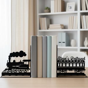 May include: Black metal train bookends with a steam engine and a train car design. The bookends are holding a stack of books. The text "Steve's Railroad" is on one bookend. The text "CraftName sign" is at the bottom.