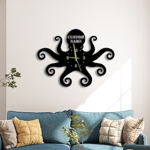 Op de afbeelding: Zwarte octopusvormige wandklok met de tekst "CUSTOM NAME" boven de wijzerplaat. De klok heeft gestileerde tentakels en een minimalistisch ontwerp. De wijzers van de klok zijn lichtgroen. De klok hangt aan een witte muur boven een blauwe bank.