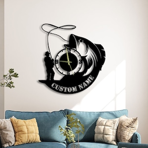 May include: Black and white wall clock with a silhouette of a fisherman casting a line at a fish. The clock face is round and has black Roman numeral markings. The clock is mounted on a white wall with a couch and a plant in the foreground. The clock has the text "CUSTOM NAME" on the bottom.