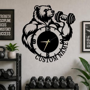 Puede incluir: Reloj de pared de metal negro con un oso levantando una mancuerna, con una esfera en el centro. El reloj tiene manecillas doradas y el texto "CUSTOM NAME" en la parte inferior. La imagen también incluye el texto "STRENGTH DISCIPLINE FOCUS CONSISTENCY SUCCESS".