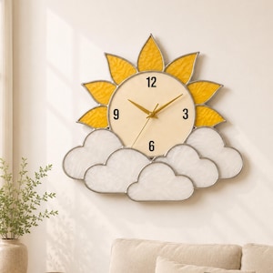 May include: A decorative wall clock shaped like a sun with yellow rays and a cream-colored clock face. The clock is surrounded by white cloud-shaped accents. The clock hands and the numbers 3, 6, 9, and 12 are visible.