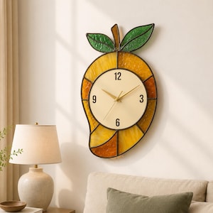May include: A mango-shaped wall clock with a stained-glass design. The clock face is cream-colored with gold hands and black numerals. The mango is yellow, orange, and brown with green leaves. The clock is hanging on a white wall.