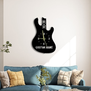 Könnte beinhalten: Schwarze Wanduhr in Form einer E-Gitarre mit weißem Zifferblatt und schwarzen Zeigern. Das Zifferblatt trägt den Text "AC/DC" und "CUSTOM NAME".