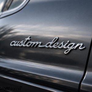 Könnte beinhalten: Eine dunkelgraue metallische Oberfläche mit dem Schriftzug custom design in erhabener, chromfarbener Schreibschrift. Der Schriftzug ist auf der glatten, reflektierenden Platte zentriert, die subtile Lichtreflexe und Schatten aufweist.