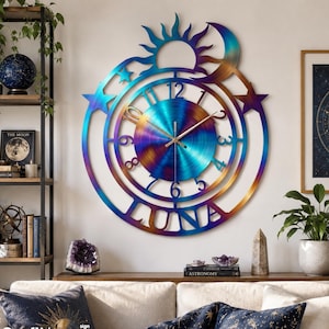 May include: A circular metal wall clock featuring a heat-treated iridescent finish with blue, purple, and gold tones. The clock face displays numbers 1 through 12, with a sun, moon, and star cutout design at the top and the word LUNA at the bottom.