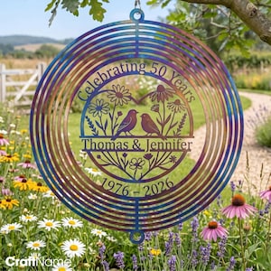 May include: A circular metal wind spinner with a heat-treated iridescent finish. The center features a heart-shaped cutout with two birds, flowers, and the text: Celebrating 50 Years, Thomas & Jennifer, and the years 1976 - 2026.