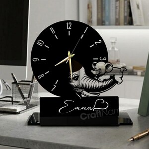Può includere: Un orologio da scrivania nero con quadrante circolare e lancette dorate. L'orologio presenta un design decorativo a forma di topo e il nome "Emma" in corsivo. L'orologio è esposto su una scrivania con libri e una penna.