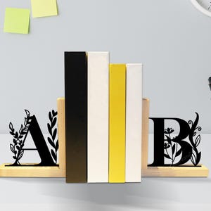 May include: Wooden bookends with black, floral-designed letters "A" and "B". The bookends hold a stack of books in black, white, and yellow. The CraftName sign is visible in the top left corner.