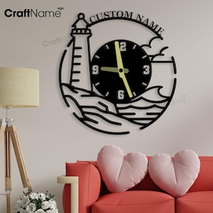 Puede incluir: Reloj de pared de metal negro con forma de faro, con esfera negra y manecillas verde neón. El reloj está rodeado por un diseño circular con las palabras "CUSTOM NAME" en la parte superior. El reloj está montado en una pared blanca sobre un sofá rojo.