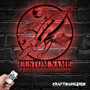 Op de afbeelding: Een rood verlicht metalen bord met een raket en planeten in een cirkel. Het bord heeft ruimte voor een aangepaste naam, "CUSTOM NAME", onder de raket. Het bord is op een bakstenen muur.