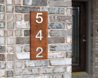Puede incluir: Un letrero rectangular vertical para número de casa con un acabado texturizado de color óxido desgastado. El letrero presenta los números metálicos grandes de color plateado 5, 4 y 2 dispuestos verticalmente en una fuente sans-serif clara.
