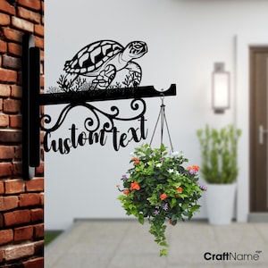 May include: Black metal wall-mounted planter with a decorative scroll design and a turtle silhouette. The planter has a hanging hook and the text "custom text" is displayed on the scroll.