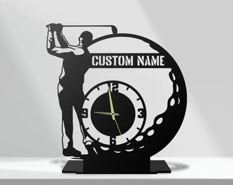 May include: A black metal desk clock featuring a silhouette of a golfer mid-swing integrated into a golf ball design. The clock face has white numerals at 3, 6, 9, and 12, with gold hands. A horizontal bar displays the text CUSTOM NAME.