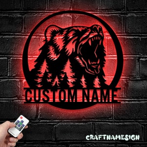 May include: A black metal silhouette of a bear's head with a roaring expression, surrounded by pine trees, in a circular frame with a red LED glow. The text "CUSTOM NAME" is displayed below the bear.