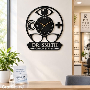 May include: A black circular wall clock featuring an eye icon, a magnifying glass, a plus sign, and eyeglasses. The clock face has gold hands and white numbers. Below the clock, a black plaque displays the text DR. SMITH OPTOMETRIST.