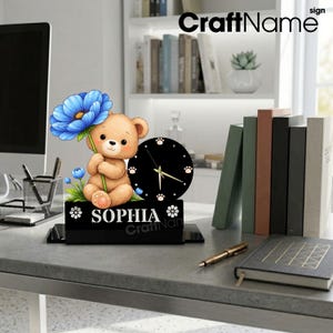 May include: A desk clock featuring a light brown teddy bear holding a large blue flower. The clock face is black with white paw print hour markers and gold hands. A black rectangular base displays the name SOPHIA in white capital letters.