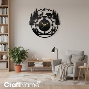 May include: A circular black wall clock featuring a laser-cut silhouette of a forest landscape with a mountain range, a howling wolf, a bear, and a deer. The clock face has a black center with gold-colored hands and large numerals.