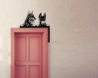 May include: A black metal door corner topper featuring two Doberman dogs peeking over the top edge. The design shows the dogs with their front paws resting on the frame, positioned at the upper right corner of a pink door.