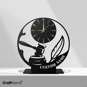 May include: A circular black metal desk clock featuring a cutout design of a judge's gavel and a quill pen. The clock face has white numbers and gold hands. The text CUSTOM NAME is cut out along the bottom right edge of the circular frame.