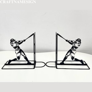 May include: Black metal bookends shaped like baseball players swinging bats. The bookends are on a white surface.