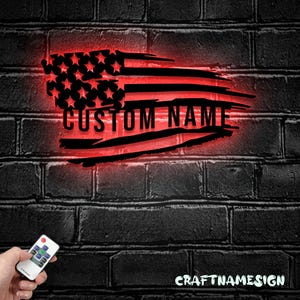 May include: A black metal wall sign shaped like a distressed American flag with a red LED backlight. The sign features a cutout star field and horizontal stripes with the text CUSTOM NAME in the center. A small remote control is visible.