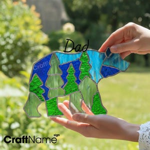 May include: A stained glass bear suncatcher featuring a geometric mosaic pattern of blue and green glass segments. The bear shape includes stylized tree silhouettes within its body. The word Dad is written in black script above the bear.