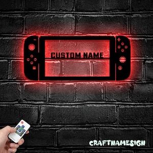 May include: A red illuminated wall sign shaped like a video game console with the text "CUSTOM NAME" displayed on the screen. The sign is mounted on a brick wall. A remote control is in the foreground.
