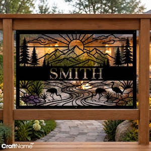 May include: A rectangular black metal sign featuring a laser-cut design of a sun rising over mountain peaks and pine trees. The center displays the name SMITH in bold capital letters. The lower section depicts a river with birds wading.
