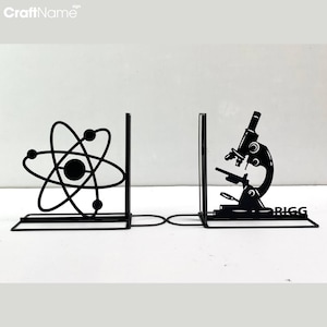 以下が含まれることがあります： 原子と顕微鏡が描かれた黒い金属製のブックエンド。ブックエンドは金属製の棒で繋がっています。右側のブックエンドには「BIGG」という文字が印刷されています。