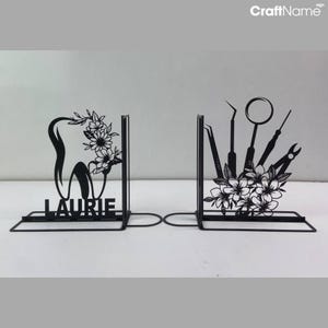 May include: Black metal bookends with a floral design. One bookend features a tooth with the name "Laurie" and the other features dental tools.