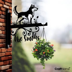 Op de afbeelding: Zwart metalen hangbord met een husky-hondsilhouet, gras en de familienaam "The Smiths". Een hangmand met kleurrijke bloemen is eronder opgehangen. Het bord is aan een bakstenen muur bevestigd.