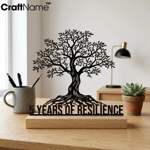 May include: A black metal tree silhouette mounted on a rectangular light wood base. The base features the text 5 YEARS OF RESILIENCE in black capital letters. The tree has intricate branches and exposed roots, standing upright as a decorative piece.