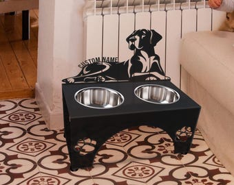 May include: A black metal elevated pet feeder with two stainless steel bowls. The stand features a cutout silhouette of a dog lying down with the text CUSTOM NAME above it and paw print cutouts on the side panels of the base structure.