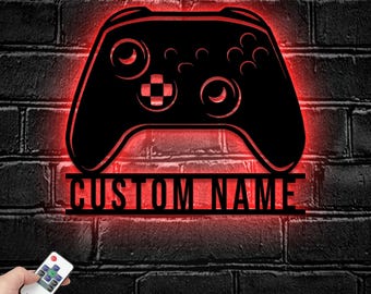 May include: A black wall-mounted sign shaped like a video game controller with a bright red LED backlight. The sign features the text CUSTOM NAME in bold black letters. A small remote control with various buttons is visible in the lower corner.