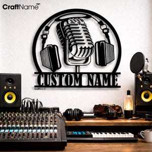 May include: A black metal wall sign featuring a circular frame containing a vintage-style microphone and headphones. Below the circular design, a horizontal bar displays the text CUSTOM NAME in a bold, capitalized, sans-serif font.