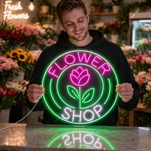 May include: A round neon sign with the words "FLOWER SHOP" in white, outlined in green, and a pink flower in the center. The sign is held by a person in front of a flower shop. The words "Fresh Flowers" are in white neon in the background.