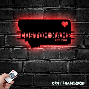 Pode incluir: Uma silhueta de metal preto do estado de Montana com um coração vermelho e o texto "CUSTOM NAME EST. 2011" iluminado em vermelho. A silhueta está montada em uma parede de tijolos.