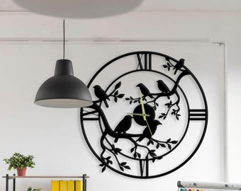 May include: A large, circular black metal wall clock featuring a cutout design of birds perched on tree branches. The clock face includes Roman numerals at the twelve, three, six, and nine o'clock positions, with thin, light-colored clock hands.