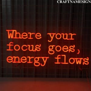 May include: Neon sign with the text "Where your focus goes, energy flows" in bright orange letters against a dark background.