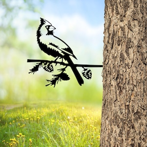 May include: Black metal silhouette of a cardinal perched on a branch with pine cones. The silhouette is attached to a tree trunk.