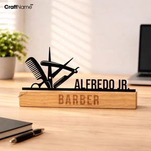 Puede incluir: Un letrero de barbero de madera con el nombre "ALFREDO JR." y la palabra "BARBER" grabados en la madera. Una silueta metálica negra de herramientas de barbero, que incluye tijeras, un peine y una navaja, se muestra encima del nombre.