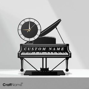 May include: A black wall clock shaped like a grand piano with a circular clock face featuring white numerals and gold hands. The piano body includes a keyboard graphic and the text CUSTOM NAME in white capital letters on the front panel.