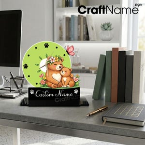 May include: A round desk clock with a light green face featuring a cartoon illustration of a mother bear and cub wearing flower crowns, a pink butterfly, and black paw prints. The black base displays the text Custom Name in white script.