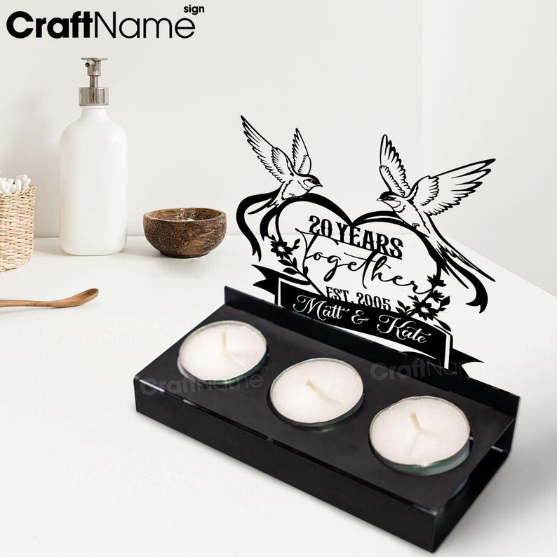 Puede incluir: Un portavelas de metal negro con tres velas de t&eacute; encendidas. Encima de las velas hay un letrero negro con dos p&aacute;jaros y el texto "20 Years Together Est. 2005 Matt & Kate" en forma de coraz&oacute;n.