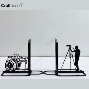 Puede incluir: Sujetalibros de metal negro con diseños de cámara y silueta de fotógrafo. Los sujetalibros tienen una base rectangular y un soporte vertical. Uno presenta una cámara detallada, el otro un fotógrafo con trípode.