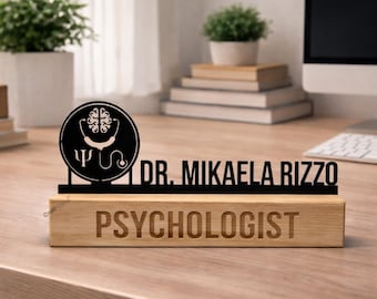 May include: A black desk sign featuring a circular icon with a brain and stethoscope, the text DR. MIKAELA RIZZO, and a light wood base with the word PSYCHOLOGIST engraved in a bold, sans-serif font, displayed on a wooden office desk.