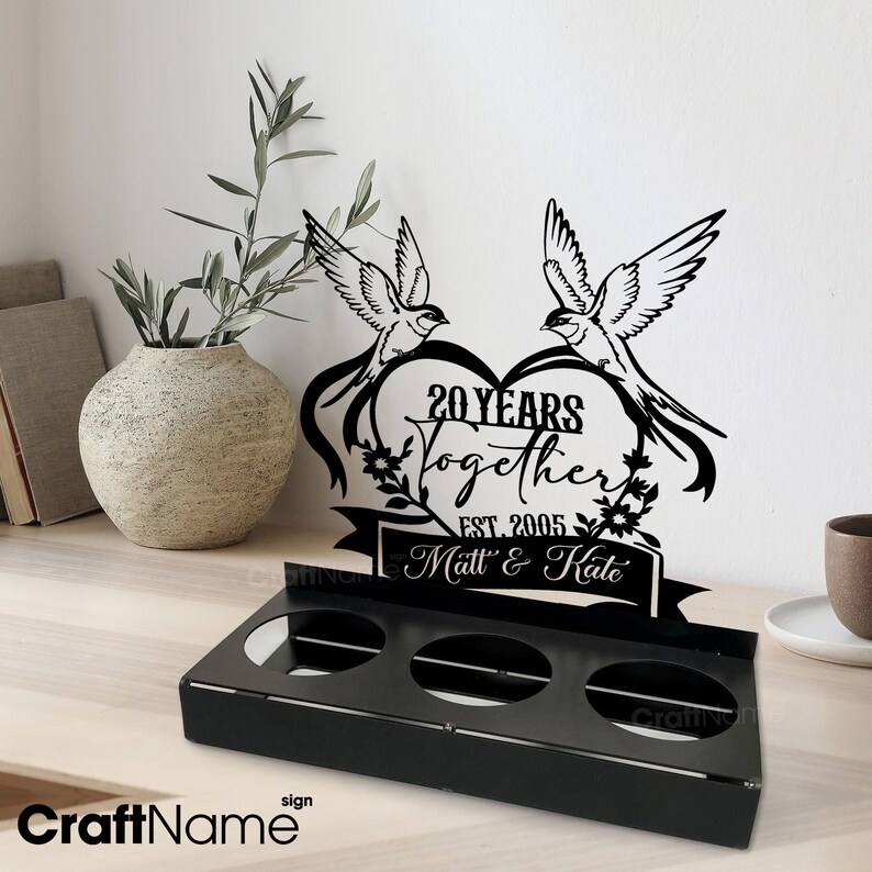 Puede incluir: Letrero de metal negro con tres aberturas circulares, con un dise&ntilde;o de coraz&oacute;n con dos p&aacute;jaros y el texto "20 Years Together Est. 2005 Matt & Kate". Un jarr&oacute;n decorativo con vegetaci&oacute;n est&aacute; en el fondo.